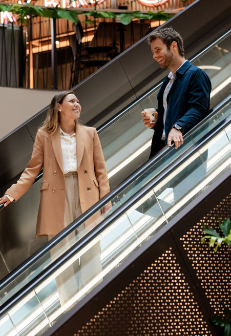 A young man and woman in professional clothes talk on escalator in a shopping center.